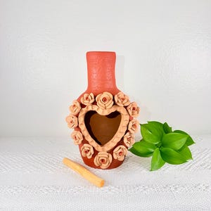 May include: A terracotta-colored vase with a heart-shaped cutout, adorned with peach-colored rose accents. A piece of palo santo wood and green leaves are next to the vase. The vase is on a white patterned surface.