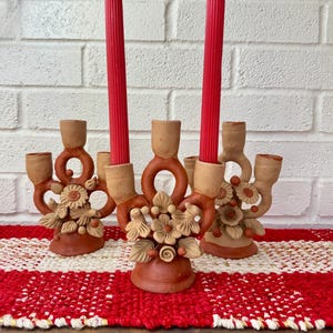 May include: Three terracotta candle holders with floral designs and red candles. The candle holders are arranged on a red and white checkered runner. The background is a white brick wall.
