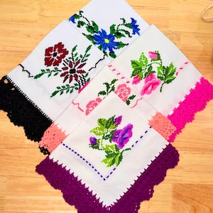May include: White embroidered handkerchiefs with floral designs in various colors, including red, blue, pink, and purple. Each handkerchief features a crocheted lace border in black, pink, or purple. The handkerchiefs are arranged on a wooden surface.