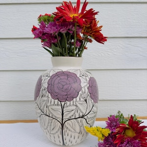 May include: A white ceramic vase with a floral design in black and purple. The vase is filled with a bouquet of red, purple, and yellow flowers.