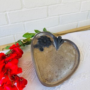 以下が含まれることがあります： 茶色の釉薬と黒い花柄が施されたハート型の陶器の皿。皿は白い表面の上に置かれている。