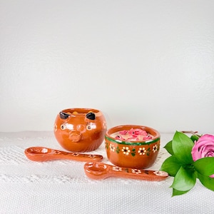 May include: Hand-painted terracotta pottery set featuring a pig-shaped bowl, a round bowl with floral accents, and two small spoons. The bowls are filled with pink items. The set is displayed on a white cloth.