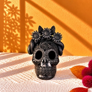 May include: A matte black skull figurine featuring intricate floral carvings across the forehead and eye sockets, topped with a sculpted crown of three large sunflowers and leaves. The skull has hollow eye sockets and a detailed jawline design.