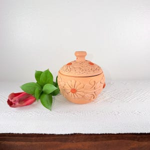May include: A terracotta-colored ceramic jar with a lid, adorned with floral relief carvings and orange accents. The jar is round with a small handle on top. A red tulip and green leaves are next to the jar.