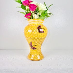 May include: A yellow ceramic vase with a butterfly design, filled with pink and white roses and green leaves. The vase has a patterned design and a decorative rim. The vase is a decorative item for the home.