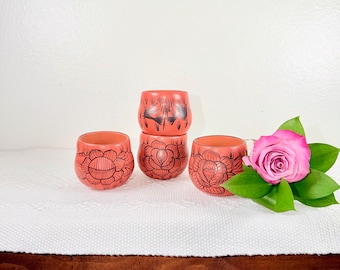 May include: Four terracotta-colored ceramic cups with black floral designs. The cups are arranged in a group, with a pink rose and green leaves to the right. The cups have a rounded shape and are likely for drinking.