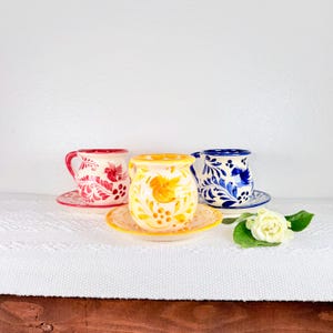 May include: Three hand-painted ceramic cups and saucers. The cups are in red, yellow, and blue, each with a unique floral design. The cups are on a white surface with a white flower and green leaves.