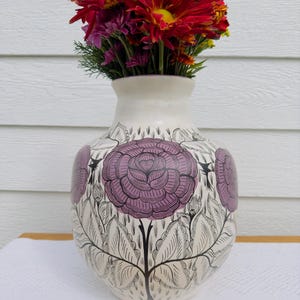 May include: A white ceramic vase with a floral design featuring purple roses and black leaves. The vase is filled with a bouquet of red and purple flowers.