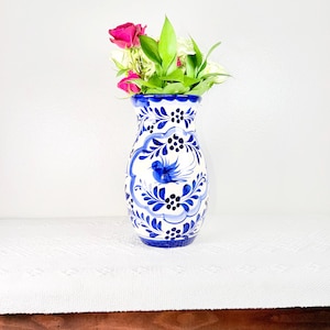 May include: A white ceramic vase featuring hand-painted cobalt blue floral patterns and a central bird motif. The vase has a flared rim and a rounded body, currently holding a bouquet of fresh pink and white roses with green leaves.