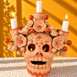 May include: A terracotta skull candle holder featuring three white taper candles. The skull is decorated with sculpted clay flowers, small birds, and round red accents. The surface of the skull has carved floral patterns and a wavy red line at the base.