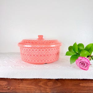 May include: A coral-colored ceramic casserole dish with a lid, adorned with a white floral pattern. The dish sits on a white cloth, with a pink rose and green leaves to the right. The dish has a small handle on the lid.