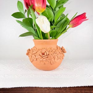 May include: A terracotta vase with floral relief holds a bouquet of red and white tulips and green foliage. The vase has a round shape with detailed flower and leaf carvings. The flowers are arranged against a white background.