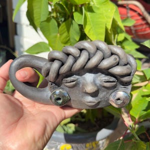 May include: A handmade ceramic mug with a face design. The mug is gray with a braided hair design on top and two green-colored eye accents. The mug is held in a hand, with green foliage in the background.
