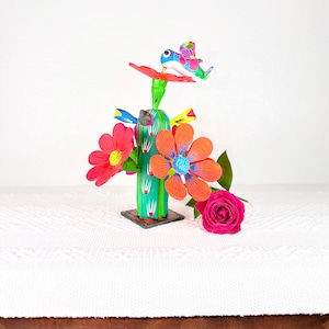May include: A colorful, handcrafted cactus sculpture with vibrant red, orange, and blue flowers. A whimsical bird perches atop the cactus, adding a playful touch. The sculpture is set against a white background, with a pink rose in the foreground.