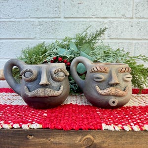 May include: Two gray ceramic mugs with faces, each featuring a handle, a nose, a mustache, and closed or open eyes with eyelashes. The mugs are displayed on a red and white patterned surface, with greenery and red berries in the background.