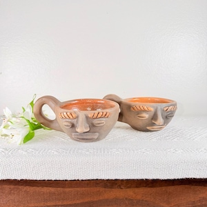 May include: Two handmade ceramic mugs with faces, each featuring a handle and an orange interior. The mugs are gray with carved facial features, including closed eyes and a defined nose and mouth. They sit on a white textured surface.