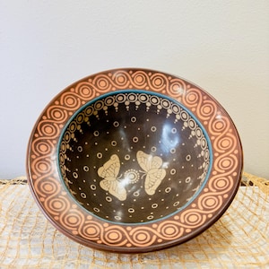 May include: A brown ceramic bowl with a decorative design featuring two butterflies and a border of orange and blue swirls. The bowl is decorated with white dots.