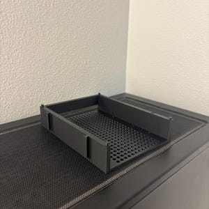 May include: A black rectangular plastic tray featuring a perforated base with a grid of small circular holes. The tray has three solid vertical walls and small protruding tabs along the sides, designed for modular organization or storage.