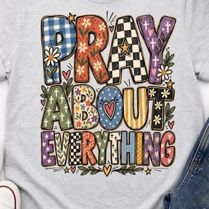 Może przedstawiać: Jasnoszary t-shirt z kolorowym nadrukiem graficznym z napisem PRAY ABOUT EVERYTHING. Litery ozdobione są różnymi wzorami, w tym niebieską kratką, motywami kwiatowymi, czarno-białą szachownicą oraz małymi krzyżykami.