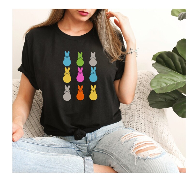 May include: A black t-shirt featuring a colourful design of nine Easter bunnies wearing glasses. The bunnies are in various colours, including blue, green, pink, yellow, orange, and grey.