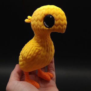 May include: A small, yellow, 3D printed bird with large black eyes and a textured surface. The bird is standing on a hand.