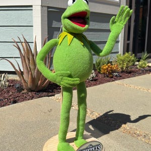 以下が含まれることがあります： 木製の台座に立っている、緑色のぬいぐるみカーミット・ザ・フロッグの人形。カエルは大きく口を開け、白い目と黄色の襟をしている。人形は片手で手を振っている。台座の看板には「The Muppets」と書かれている。