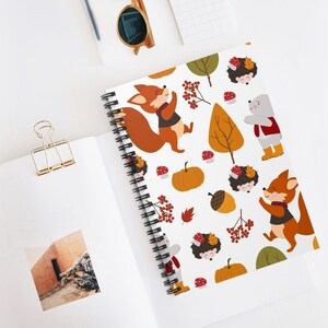 Puede incluir: Cuaderno de espiral con un patrón otoñal. El diseño incluye zorros, erizos, calabazas, bellotas y hojas de otoño en tonos naranja, marrón y rojo sobre un fondo blanco. El cuaderno tiene una encuadernación en espiral negra.