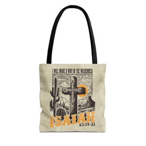 May include: Beige tote bag with black handles. The design features a desert landscape with a cross, cactus, and the text "I WILL MAKE A WAY IN THE WILDERNESS" and "ISAIAH 43:19-21" in orange.