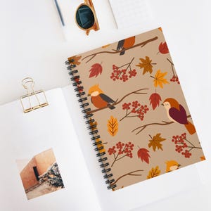 May include: Spiral-bound notebook featuring a fall-themed pattern. The design includes illustrations of birds perched on branches, along with leaves and berries in shades of orange, red, and brown against a tan background. The notebook is open, revealing blank pages.
