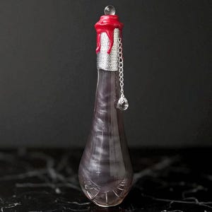 May include: A decorative glass bottle with a pearlescent purple liquid. The bottle is sealed with a red wax top and a silver band. A silver chain with a crystal pendant hangs from the bottle.