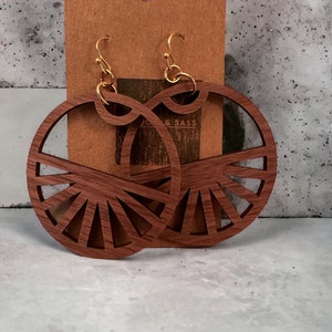 May include: A pair of large, round, wooden earrings with a sunburst design. The earrings are brown and have gold hooks.
