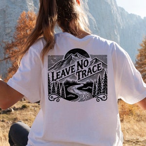 May include: A white t-shirt featuring a black graphic print of a mountain range, a river, and trees. The text "LEAVE NO TRACE" is printed in a vintage style font.