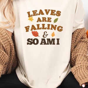 May include: Cream-colored t-shirt with the text "LEAVES ARE FALLING & SO AM I" in brown and orange lettering. The shirt features leaf graphics. The person is wearing a brown knitted cardigan and black pants.