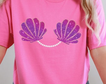 May include: Pink t-shirt featuring a purple glitter seashell design and a pearl necklace.