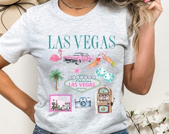 May include: Grey t-shirt with a graphic design featuring the text "LAS VEGAS" and various images related to Las Vegas, including a pink flamingo, a pink car, a palm tree, dice, a slot machine, and a camera.