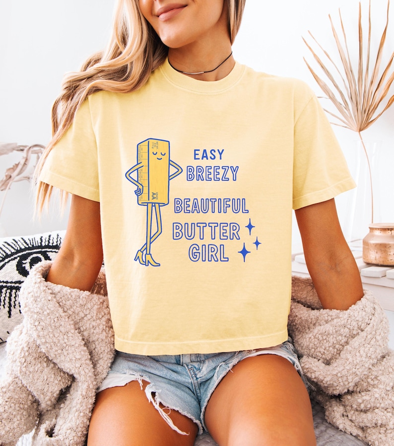 May include: A pale yellow t-shirt featuring the words "EASY BREEZY BEAUTIFUL BUTTER GIRL" and a cartoon butter stick character. The butter stick has arms, legs, and is wearing high heels. The shirt is a casual style.
