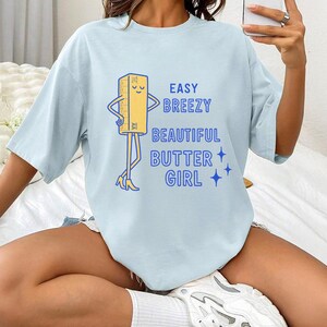 May include: Pale blue t-shirt featuring the text "EASY BREEZY BEAUTIFUL BUTTER GIRL" and a cartoon butter stick character. The butter stick has arms, legs, and is wearing high heels. The design is in blue and yellow.