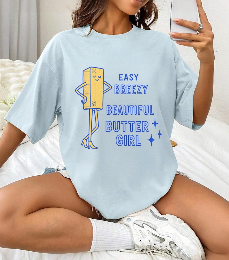 May include: Pale blue t-shirt featuring the text "EASY BREEZY BEAUTIFUL BUTTER GIRL" and a cartoon butter stick character. The butter stick has arms, legs, and is wearing high heels. The design is in blue and yellow.
