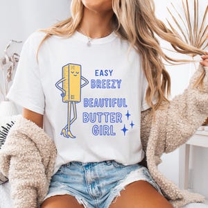 May include: White t-shirt with the text "EASY BREEZY BEAUTIFUL BUTTER GIRL" in blue. The shirt features a cartoon butter stick character with arms, legs, and high heels. The butter stick is yellow with blue accents.