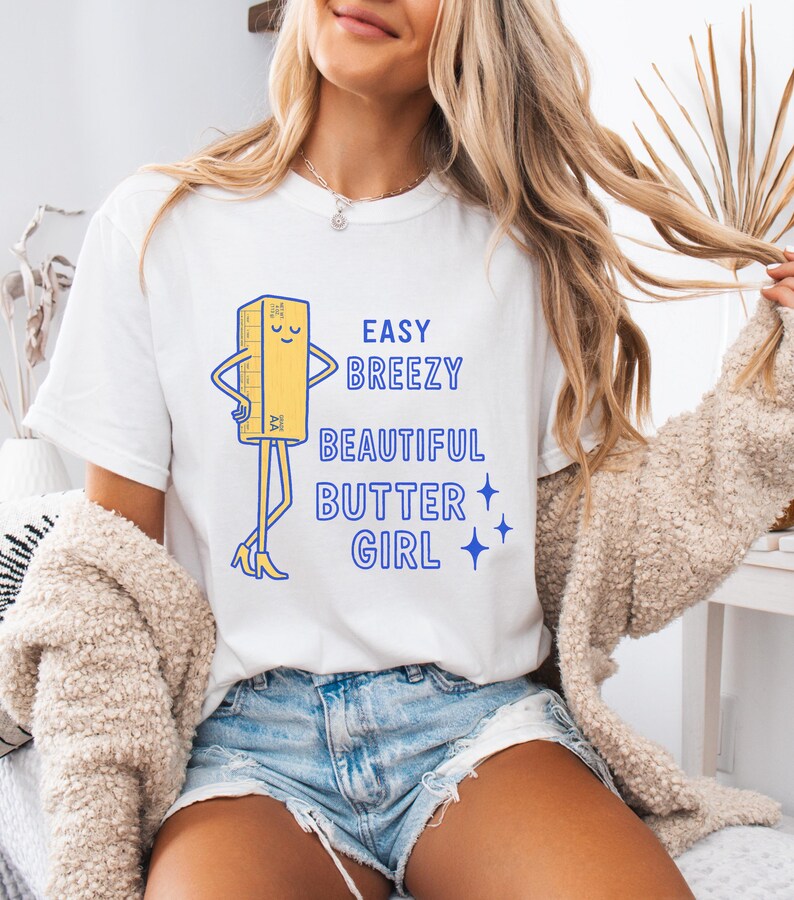 May include: White t-shirt with the text "EASY BREEZY BEAUTIFUL BUTTER GIRL" in blue. The shirt features a cartoon butter stick character with arms, legs, and high heels. The butter stick is yellow with blue accents.