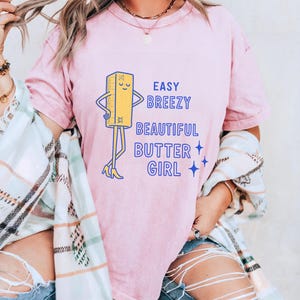 May include: A pale pink t-shirt with the slogan "Easy Breezy Beautiful Butter Girl" in blue. The design includes a cartoon butter stick character with arms, legs, and high heels. The t-shirt has a relaxed fit.
