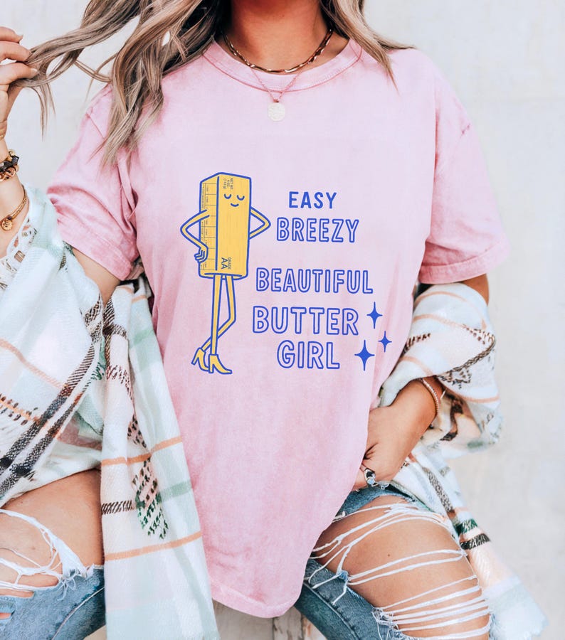 May include: A pale pink t-shirt with the slogan "Easy Breezy Beautiful Butter Girl" in blue. The design includes a cartoon butter stick character with arms, legs, and high heels. The t-shirt has a relaxed fit.