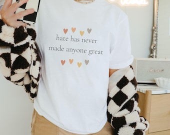 Op de afbeelding: Wit T-shirt met de tekst "hate has never made anyone great" in zwart gedrukt. De tekst wordt omringd door kleine, kleurrijke hartjes.