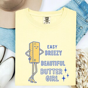 May include: A pale yellow sweatshirt featuring a cartoon butter stick illustration and the text "Easy Breezy Beautiful Butter Girl" in blue. The sweatshirt has a crew neck and long sleeves.