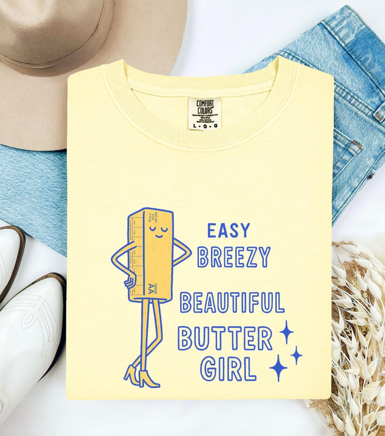 May include: A pale yellow sweatshirt featuring a cartoon butter stick illustration and the text "Easy Breezy Beautiful Butter Girl" in blue. The sweatshirt has a crew neck and long sleeves.