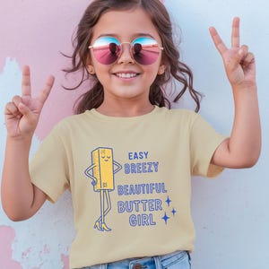 May include: A young person wearing a pale yellow t-shirt with the text "EASY BREEZY BEAUTIFUL BUTTER GIRL" and a cartoon butter stick illustration. The person is wearing round sunglasses and making a peace sign.