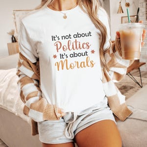 May include: White t-shirt with the text "It's not about Politics, It's about Morals" in black, pink, and gold lettering. The shirt has a crew neck and is worn with gray shorts and a brown and white striped cardigan.