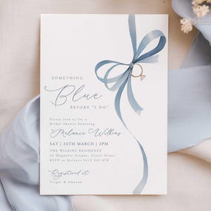 Op de afbeelding: Een witte uitnodigingskaart met de tekst "Something Blue Before I Do" in elegant schrift. Een lichtblauw lintontwerp met een gouden ring accentueert de tekst. De uitnodiging is voor Melanie Wilkins' bruidsdouche.