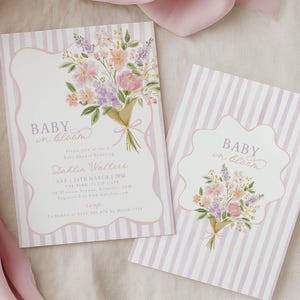 May include: Two baby shower invitations with floral bouquets and the text "Baby in Bloom." The invitations have a striped background in white and light purple. A pink fabric is draped in the background.