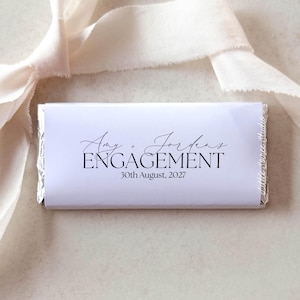 May include: A rectangular chocolate bar wrapped in white paper with black text that reads Amy + Jordan's ENGAGEMENT 30th August, 2027. The bar is accented by a long, flowing cream-coloured fabric ribbon tied in a loose knot above the item.